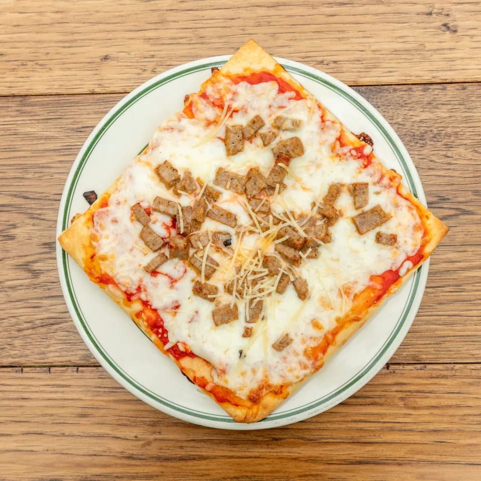 Square slice of pizza with melted cheese and sausage crumbles on a white plate with a green rim.