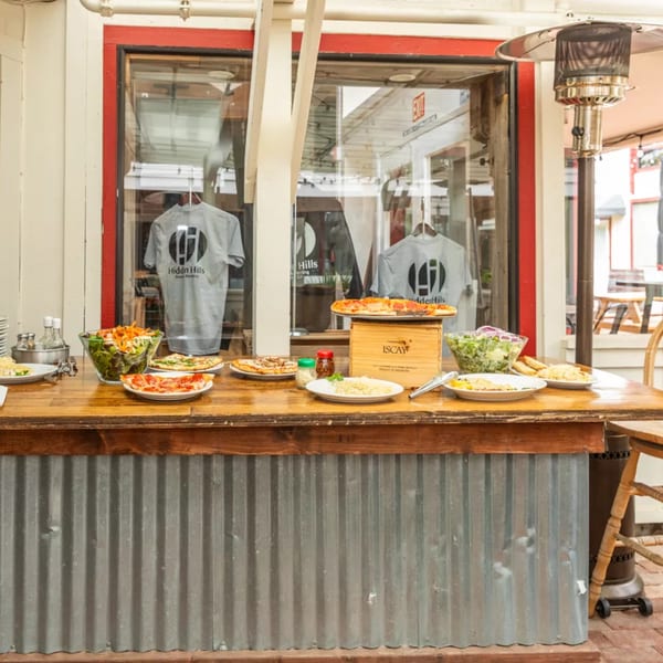 Outdoor buffet table with pizzas, salads, and pasta in front of a restaurant window.