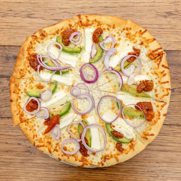 Round pizza with white cheese, avocado slices, red onion rings, and sun-dried tomatoes on wood.