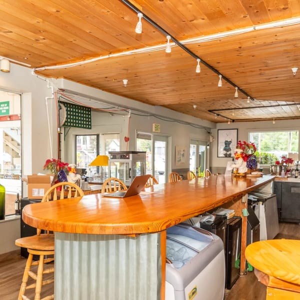 A long, polished wooden bar with stools, a laptop, and decorative roosters in a bright, rustic restaurant.