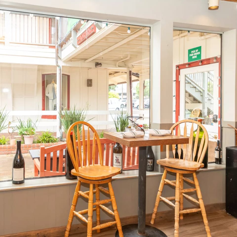 A cozy restaurant window table set for two with wine bottles, overlooking an outdoor patio.