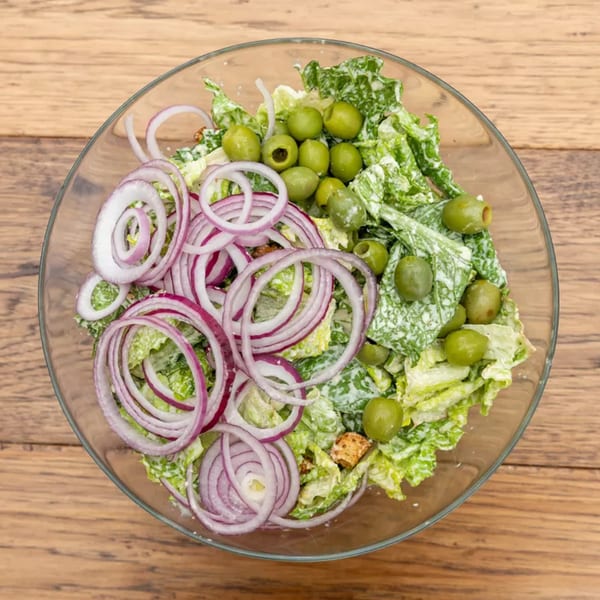 Overhead view of a Caesar salad with red onion rings and green olives in a glass bowl.