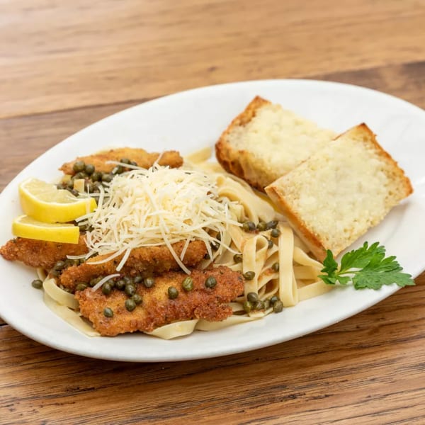 Chicken piccata with capers, lemon slices, and shredded cheese, served with pasta and garlic bread.