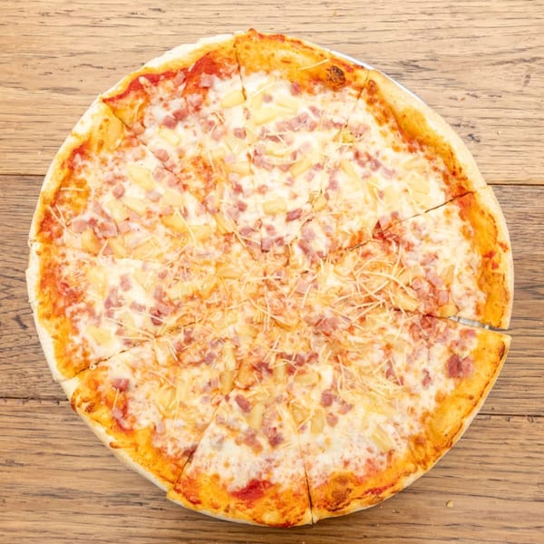 A whole Hawaiian pizza, cut into slices, with ham, pineapple, and cheese on a wooden table.